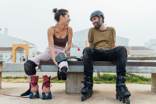 Cheerful girlfriend looking at boyfriend with roller skates while sitting on bench