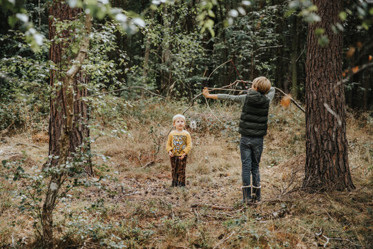 Boy playing with bow and arrow while brother standing by him in forest