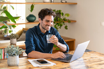 Smiling male freelance worker holding glass of water while working on laptop at home office