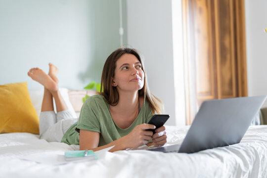 Thoughtful young woman with smart phone lying in front of laptop on bed at home