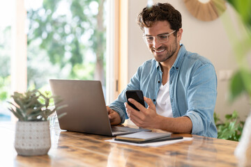 Smiling male freelancer with eyeglasses using smart phone while sitting with laptop at home office