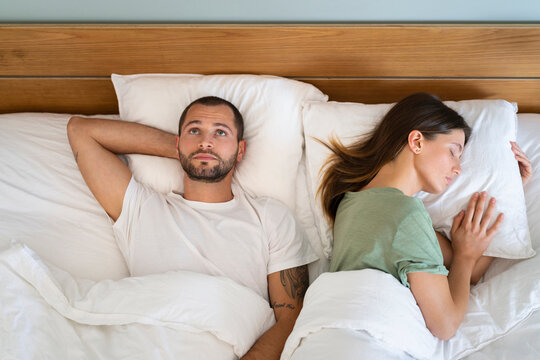 Young Woman Sleeping By Worried Man Staring At Ceiling While Lying On Bed At Home