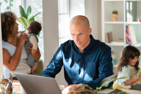 Father working on laptop while family running in background at home office