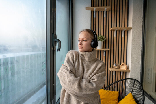 Young woman listening music through headphones while standing with arms crossed by window