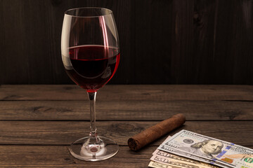 Glass of red wine with money and cuban cigar on an old wooden table. Focus on the cuban cigar