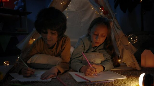 Joyful boy and girl drawing with colored pencils while spending free time in evening. Children lying on floor in decorative tent while creating pictures. Concept of leisure.