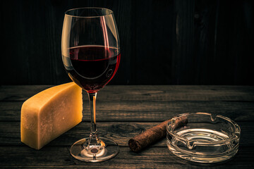 Glass of red wine with a piece of parmesan and cuban cigar with glass ashtray on an old wooden table. Focus on the cuban cigar