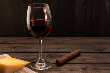 Glass of red wine with a piece of parmesan and cuban cigar on an old wooden table. Focus on the cuban cigar