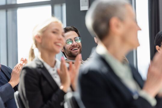 selective focus of smiling businessman near colleagues applauding on blurred foreground