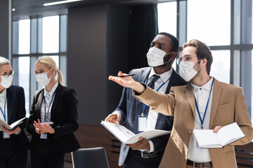 young businessman in medical mask pointing with hand during conversation with african american colleague