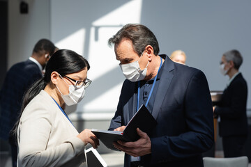 Fototapeta na wymiar mature businessman in medical mask showing notebook to asian colleague