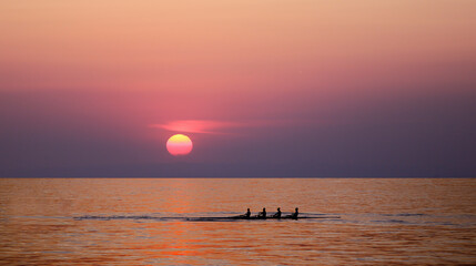 People silhouettes on rowing boat in front of sunset background. Front view - Powered by Adobe