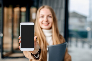 Fototapeta premium Happy smiling woman showing mobile phone against the background of the office center. Mockup for design