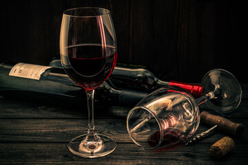 Two bottles of red wine with two glasses and corkscrew with cork on an old wooden table. Focus on an overturned glass of wine
