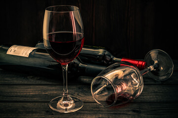 Two bottles of red wine with two glasses on an old wooden table. Focus on an overturned glass of wine