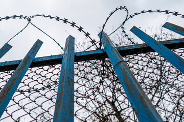 A blue fence with barbed wire near prison or mental hospital on the background of grey sky.