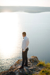 Stylish groom standing on the stone near the beautiful lake