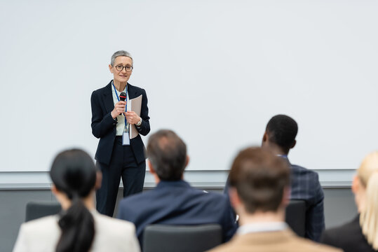 Smiling businesswoman with microphone and paper folder talking near blurred business people in conference room
