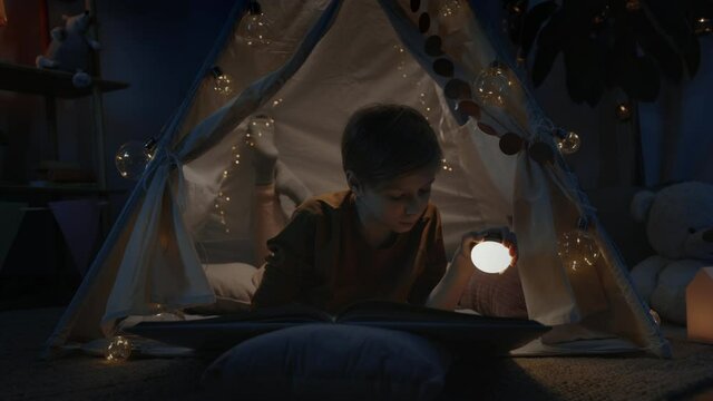 Teen boy in makeshift hut using flashlight while reading book at home in evening. Curious teenager lying on floor while spending free time. Concept of leisure and careless childhood.