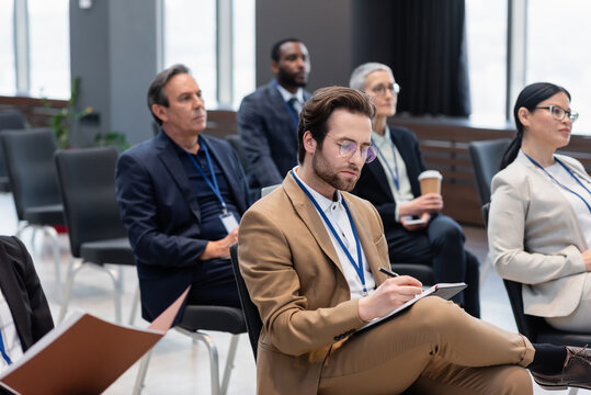 Businessman writing on notebook near blurred interracial business people in conference room
