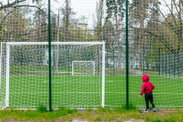 Little boy in a red jacket next to the soccer field. The kid and the soccer stadium behind the fence