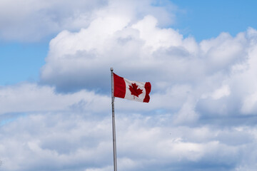 The National Flag of Canada, blowing left to right  in the wind off a flag pole against a cloud filled light blue sky background
