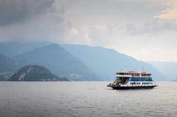 Early morning view of the Como Lake with a ferry boat sailing near the harbor of Varenna, Italy, cloudy sky, with fog, and mountains in the distance