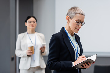 Mature businesswoman writing on notebook near asian colleague on blurred background