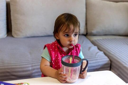 Photo of cute baby girl holding plastic glass with straw while drinking homemade milkshake. Caucasian baby girl having her milk looking away. Happy child enjoy with a glass of baby food in her hand.