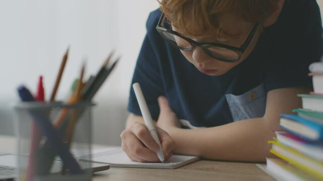 Concentrated little boy studying to write, writing letters in notebook at home workplace, close up portrait, slow motion