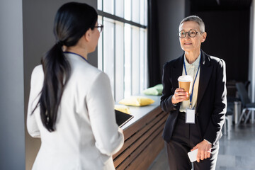 middle aged businesswoman holding takeaway drink and cellphone during conversation with blurred colleague