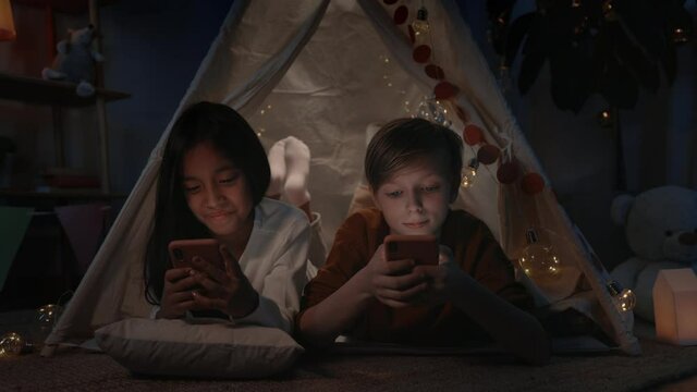 Teenagers spending time while browsing internet in decorative makeshift hut at home in evening. Girl and boy lying on floor while using smartphones.Concept of leisure.