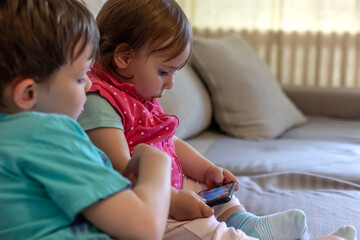 Big brother sharing funny content on smartphone with his younger sister. Two kids with gadgets. Little girl and boy sitting on sofa with a smart phone at home. Happy children playing indoors.