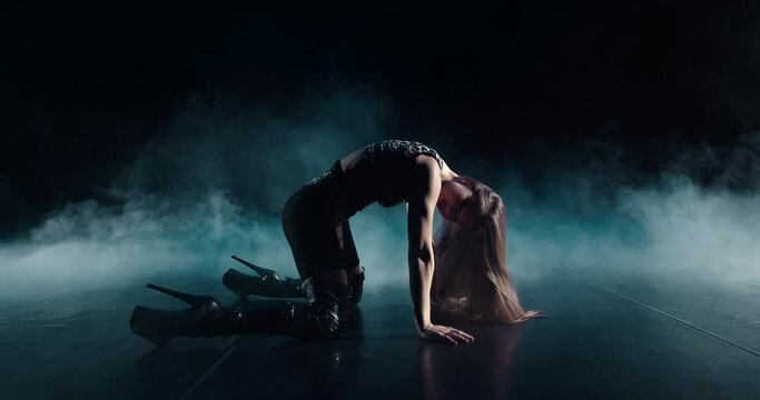 A sympathetic girl is dancing strip dances on the floor in smoke. She is wearing an attractive, sexy suit with heels on her feet. She makes erotic and fluid movements of her legs, arms and body