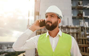 Serious male contractor speaking on smartphone at construction site