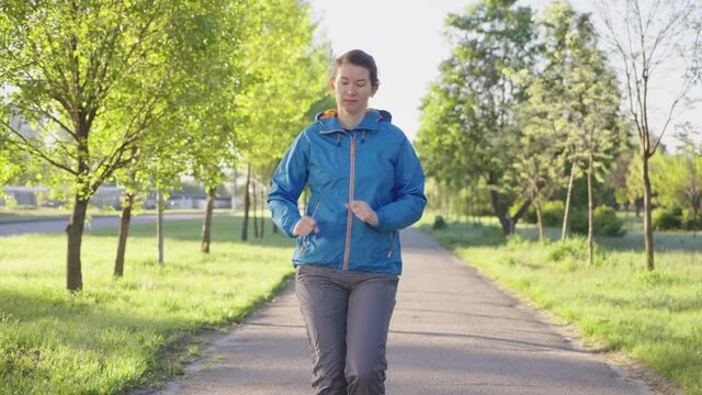 Sporty woman wearing blue jacket and gray pants walking in park in the morning, rays of sunrise on green trees. Tracking shot jogger training outside. Concept of fitness