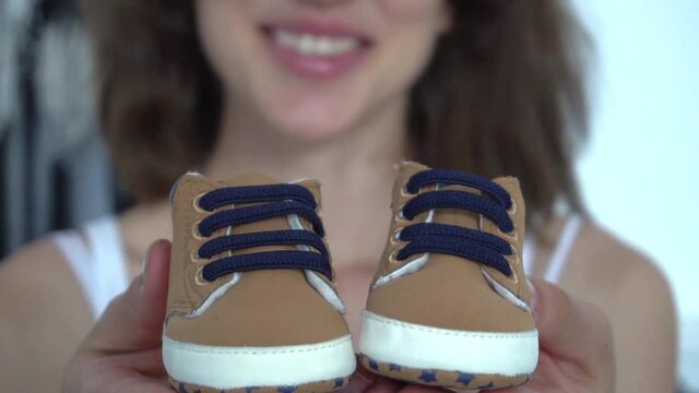 Cropped image of pregnant woman holding newborn shoes in bedroom
