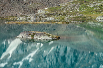 Fototapeta premium Rock in turquoise mountain lake. Snowy mountain reflected in azure clear water of glacial lake. Beautiful sunny background with snow-white glacier reflection in green water surface of mountain lake.