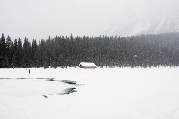 One person dressed in red in a winter landscape. Frozen lake, wooden house and a forest under the snow. Banff National Park, Alberta, Canada
