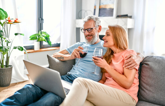 A mature couple is sitting on the couch at home with a laptop and glasses of red wine. Stay home concept. Conversation on a video call. Romantic evening for a married couple. - Powered by Adobe