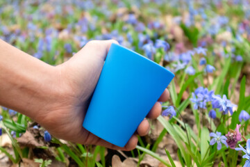 Hand holding blue reusable plastic glass for picnic in wild spring blooming lawn close-up. Travel cup in blue flowers with focus blur. Eco picnic utensil details.