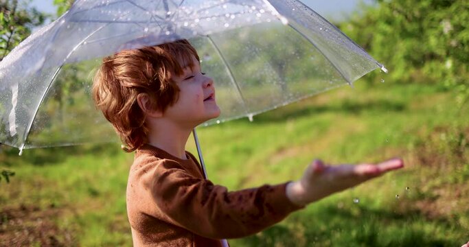 cheerful baby boy catching the rain drops, that are falling from umbrella in spring garden at sunny summer day