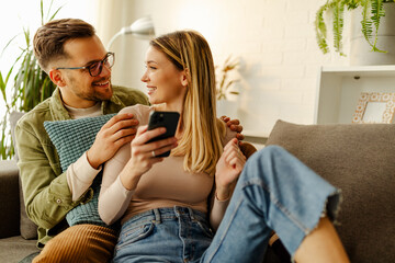 Happy young couple enjoying media content in a smart phone sitting on a couch at home.