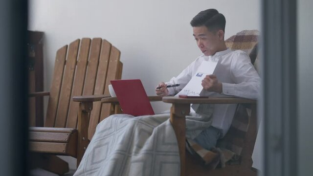 Shooting behind window of concentrated professional Asian man sitting on deck pointing at graph talking in video chat. Side view portrait of expert business consultant working in home office