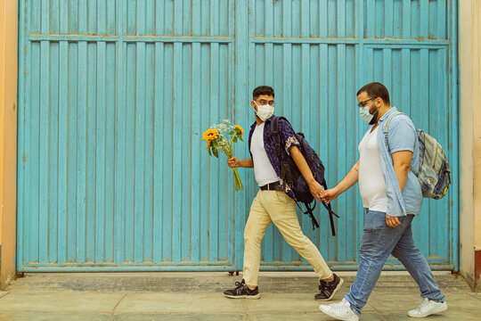 Pareja de novios gay caminando de la mano con flores en la calle usando mascarillas frente a una puerta azul 