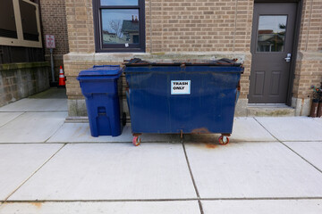 Small blue dented dumpster on wheels on a sidewalk next to a blue trash can in front of a brown brick wall