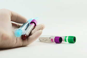 Hand with surgical glove holding vacuum tubes for collection and blood samples and a syringe on white background. Tubes with purple and green lid and label to identify the data. Selective focus.