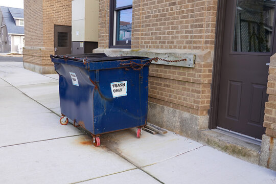 Small blue dented dumpster on wheels on a sidewalk chained to a brown brick wall