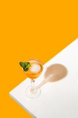 Elegant stemware glass of fresh orange cocktail with big ice ball on white table surface, bright yellow background