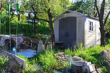 Small plastic tool storage shed placed in spring terrace garden, with round table in front, plastic bottles on stick for scaring away birds and vivid green grass around. Spring afternoon sunshine. 
